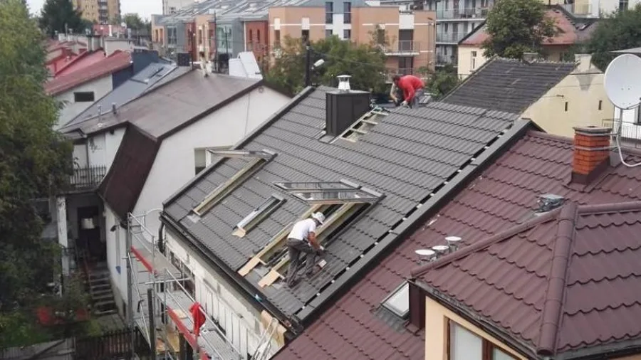 Vue plongeante sur les toits d'un quartier ancien de Cracovie : deux couvreurs posent une couverture en tuiles métalliques anthracite sur un versant.